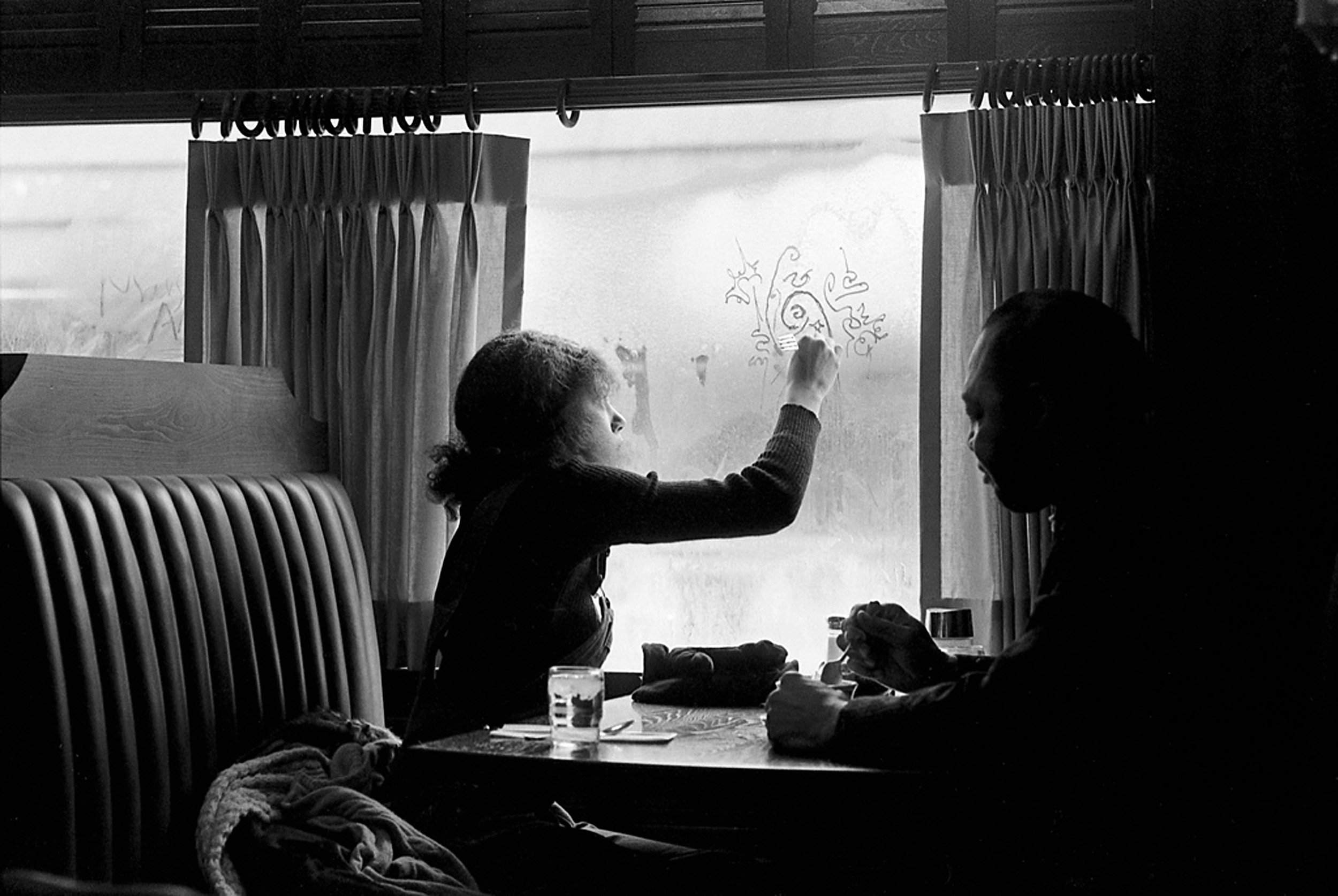 Woman in restaurant booth doodles in window fog, 1969.