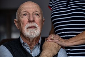 Portrait of a senior man with spouse's hand on his shoulder.